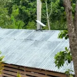 Una antena Starlink instalada sobre el techo de una casa rural, rodeada de vegetación tropical. La imagen muestra cómo esta tecnología puede integrarse en comunidades remotas para proporcionar acceso a internet de alta velocidad, destacando el contraste entre la innovación tecnológica y el entorno natural.